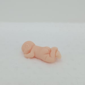 Puede incluir: Una pequeña figura de color rosa claro de un bebé acostado boca abajo. El bebé está acurrucado con la cabeza apoyada en el brazo y las piernas dobladas. La figura es lisa y detallada.