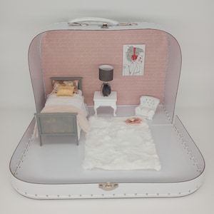 May include: A miniature dollhouse bedroom scene inside a white suitcase. The interior features a small bed, a fluffy white rug, a side table with a lamp, and a small armchair. The back wall is pink with a drawing of a woman.