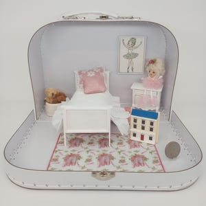 Puede incluir: Una escena de dormitorio de casa de muñecas en miniatura dentro de una maleta blanca. La escena incluye una cama con una almohada rosa, una alfombra con un patrón de zapatillas de ballet, una cómoda pequeña y un estampado de bailarina enmarcado.