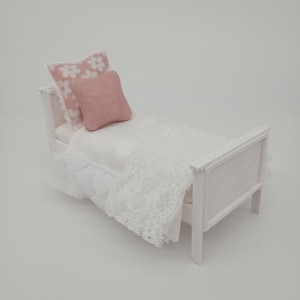 May include: A miniature white bed with a white lace blanket and two pillows. One pillow is solid pink, and the other is white with pink floral designs. The bed frame is white.