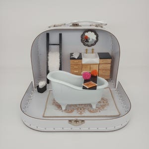 May include: A miniature bathroom scene set within a white suitcase. The scene features a white clawfoot bathtub, a small rug, a sink, a mirror, and a towel rack. Decorative items include pom-poms and a miniature book.