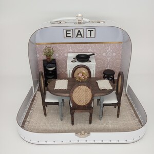 May include: A miniature dining room scene inside a white suitcase. The scene includes a table, chairs, a small stove, and a cabinet. The word "EAT" is spelled out above the scene. The interior is decorated with a patterned wallpaper and a rug.