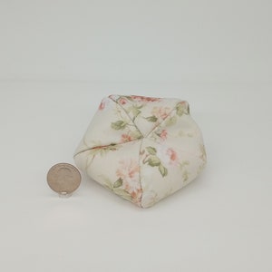 May include: A small, cream-colored, floral patterned beanbag chair. The chair is shaped like a cube with rounded edges. The floral pattern features pink roses and green leaves.