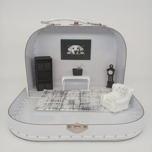 May include: A miniature white suitcase transformed into a dollhouse interior. Inside, a small rug, armchair, bookshelf, table, and a clock are arranged. A framed picture of a dog hangs on the back wall.