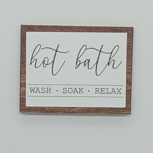 May include: A rectangular wooden sign with a white center and a brown frame. The words "hot bath" are written in cursive script above the words "WASH • SOAK • RELAX" in a simple font.