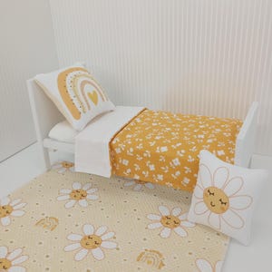 Könnte beinhalten: Ein weißes Miniaturbett mit einer gelben Bettdecke mit Blumenmuster und einem weißen Laken. Ein passendes Kissen mit Regenbogenmuster und ein Gänseblümchenkissen befinden sich auf dem Bett. Ein gelber Teppich mit Gänseblümchenmuster liegt auf dem Boden.