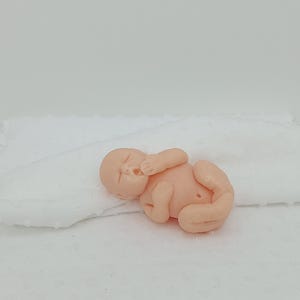 Könnte beinhalten: Eine kleine, hautfarbene Babyfigur liegt auf einer weißen, strukturierten Oberfläche. Das Baby ist zusammengerollt, die Augen geschlossen und eine Hand in der Nähe des Mundes. Die Figur ist wahrscheinlich aus einem glatten Material gefertigt.