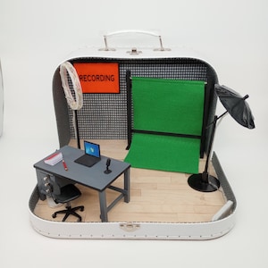 Puede incluir: Miniatura de estudio de grabación dentro de una maleta blanca. El set incluye un escritorio con un portátil, un micrófono y una silla, una pantalla verde y un telón de fondo con la palabra "RECORDING". También se incluye una pequeña luz y un paraguas.