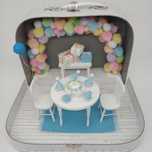 May include: A miniature birthday party scene inside a white suitcase. The scene includes a round table with a cake, party hats, and two white chairs on a blue rug. A pom-pom arch and gifts complete the festive setting.