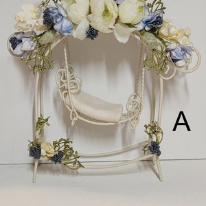 Peut inclure: Ensemble de balançoire en métal blanc avec un coussin blanc et une guirlande florale de fleurs blanches et bleues. L'ensemble de balançoire est décoré de petites baies bleues et de feuilles vertes. La lettre "A" est visible dans le coin inférieur droit de l'image.
