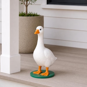 Peut inclure: Une figurine d'oie blanche avec un bec et des pattes oranges, posée sur une base circulaire verte. L'oie a un corps texturé et est placée sur une surface en bois. Un objet décoratif pour la maison ou le jardin.