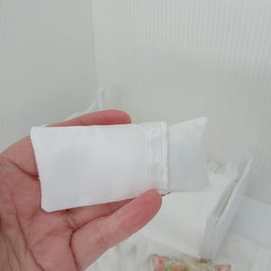 Könnte beinhalten: Ein kleines, weißes Satin-Kissen wird in einer Hand gehalten, wobei ein passendes Kissen teilweise sichtbar ist. Ein Miniaturbett und ein Blumenkissen befinden sich im Hintergrund. Die Artikel scheinen Puppenhauszubehör zu sein.
