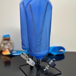 Peut inclure: Deux gourdes souples bleues translucides de 500 ml montées sur un support de séchage en plastique noir. Chaque gourde porte l'inscription 500 ml SOFTFLASK sur le côté. Le support comprend plusieurs tiges noires et une étiquette blanche Frch 15.
