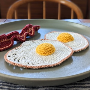 May include: Two crocheted fried eggs on a grey plate with a crocheted strip of bacon. The eggs are white with a yellow yolk and a brown border.