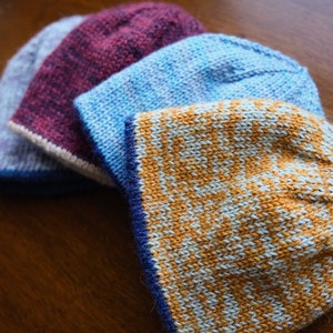 May include: Four knitted hats in different colors and patterns. The hats are stacked on top of each other. The top hat is a light blue and orange striped hat. The second hat is a light blue hat with a white and blue pattern. The third hat is a dark purple hat with a white and purple pattern. The bottom hat is a light blue hat with a white and blue pattern.