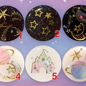 May include: Six round resin cabochons featuring various space-themed designs. The designs include planets, stars, moons, and a castle. The cabochons are all white or black with gold glitter and accents.