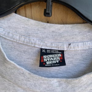Pu&ograve; includere: Primo piano di un'etichetta di una maglietta grigia XL. L'etichetta recita "50% Cotone, 50% Poliestere" e presenta il logo "Screen Stars Best" con "Made in U.S.A." stampato sotto. La maglietta &egrave; appesa a una gruccia nera.