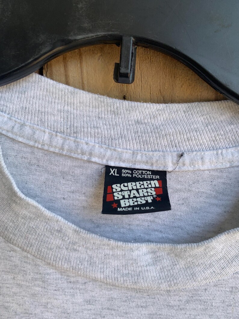 Pu&ograve; includere: Primo piano di un'etichetta di una maglietta grigia XL. L'etichetta recita "50% Cotone, 50% Poliestere" e presenta il logo "Screen Stars Best" con "Made in U.S.A." stampato sotto. La maglietta &egrave; appesa a una gruccia nera.