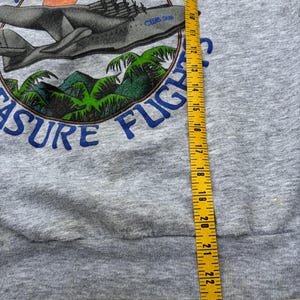 Puede incluir: Sudadera gris con un gr&aacute;fico que presenta un avi&oacute;n, palmeras y el texto "AMAZON TREASURE FLIGHT". El dise&ntilde;o incluye un emblema circular con detalles en azul y naranja. Se ve una cinta m&eacute;trica amarilla.