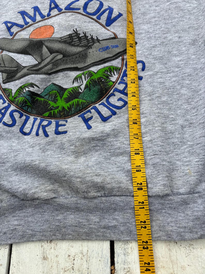 Puede incluir: Sudadera gris con un gr&aacute;fico que presenta un avi&oacute;n, palmeras y el texto "AMAZON TREASURE FLIGHT". El dise&ntilde;o incluye un emblema circular con detalles en azul y naranja. Se ve una cinta m&eacute;trica amarilla.