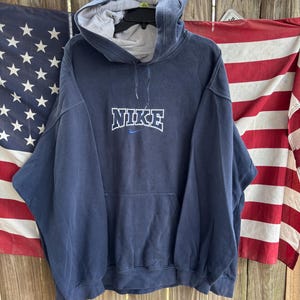 May include: Navy blue hoodie with the brand name "NIKE" in white block letters across the chest. The hoodie has a front pocket and a drawstring hood. The background includes American flags.
