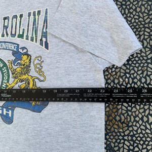 Pu&ograve; includere: T-shirt grigio chiaro con una grafica con la scritta "ROLINA" in lettere a scacchi. Sotto, una grafica di un leone e la scritta "CONFERENCE" sono visibili. La maglietta misura circa 61 cm di larghezza.