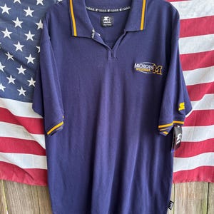 May include: Navy blue polo shirt with yellow trim on the collar and sleeves. The shirt features the text "MICHIGAN WOLVERINES" and a logo on the chest. The shirt is hanging in front of an American flag.