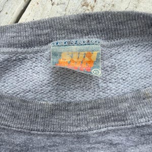 Puede incluir: Primer plano de una sudadera gris XL con cuello de canal&eacute;. La etiqueta presenta la palabra "SUN" en letras naranjas y amarillas. El fondo es una superficie de madera clara y desgastada.