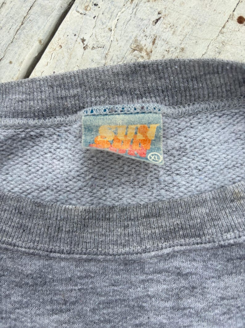 Puede incluir: Primer plano de una sudadera gris XL con cuello de canal&eacute;. La etiqueta presenta la palabra "SUN" en letras naranjas y amarillas. El fondo es una superficie de madera clara y desgastada.