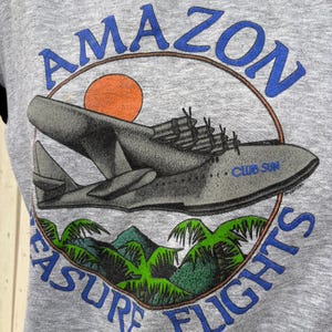 Puede incluir: Sudadera gris con un gr&aacute;fico de hidroavi&oacute;n, un sol y follaje tropical. El texto dice "AMAZON" encima del avi&oacute;n y "PLEASURE FLIGHTS" debajo. El avi&oacute;n tiene el texto "CLUB SUN".