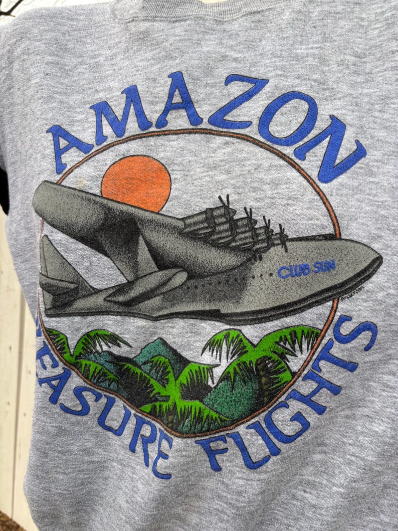 Puede incluir: Sudadera gris con un gr&aacute;fico de hidroavi&oacute;n, un sol y follaje tropical. El texto dice "AMAZON" encima del avi&oacute;n y "PLEASURE FLIGHTS" debajo. El avi&oacute;n tiene el texto "CLUB SUN".