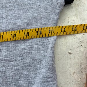 Puede incluir: Primer plano de una camiseta gris con mangas negras. Una cinta m&eacute;trica amarilla muestra una longitud de 43 a 61 cm. La tela de la camiseta tiene un aspecto texturizado.