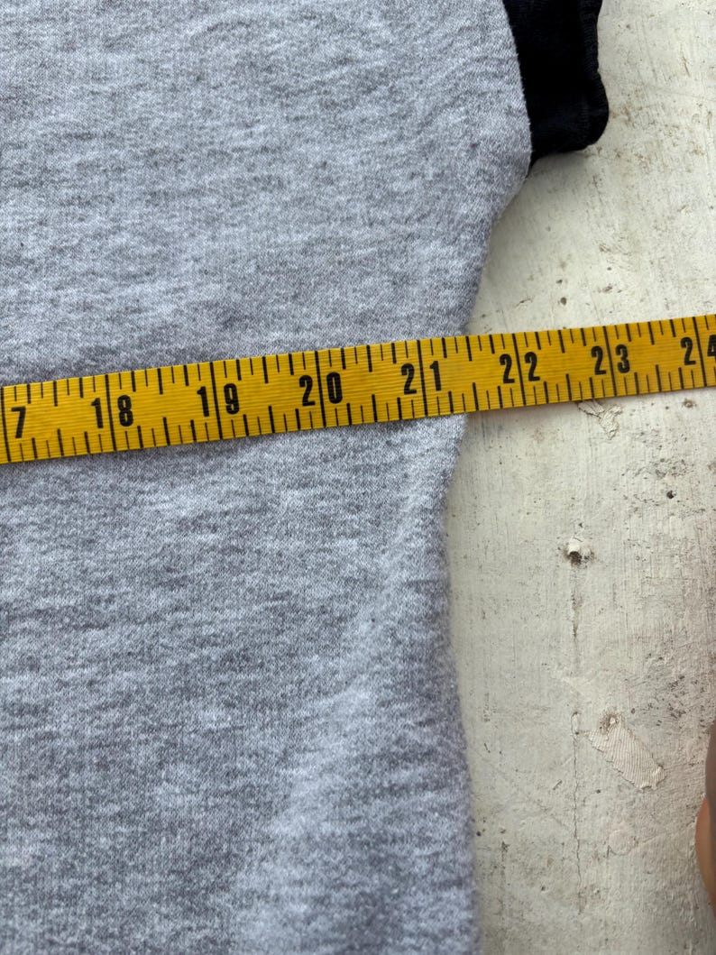 Puede incluir: Primer plano de una camiseta gris con mangas negras. Una cinta m&eacute;trica amarilla muestra una longitud de 43 a 61 cm. La tela de la camiseta tiene un aspecto texturizado.