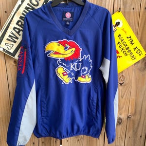 May include: A blue and gray windbreaker jacket with a large embroidered Kansas Jayhawks logo on the front. The logo features a yellow and red bird with the letters "KU" on its chest.