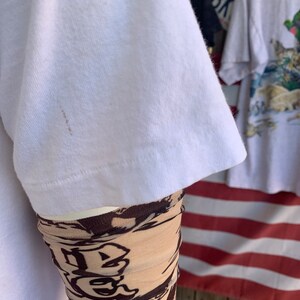 May include: White t-shirt with a brown stain on the sleeve. The sleeve is rolled up and a brown and beige patterned sleeve is visible underneath.