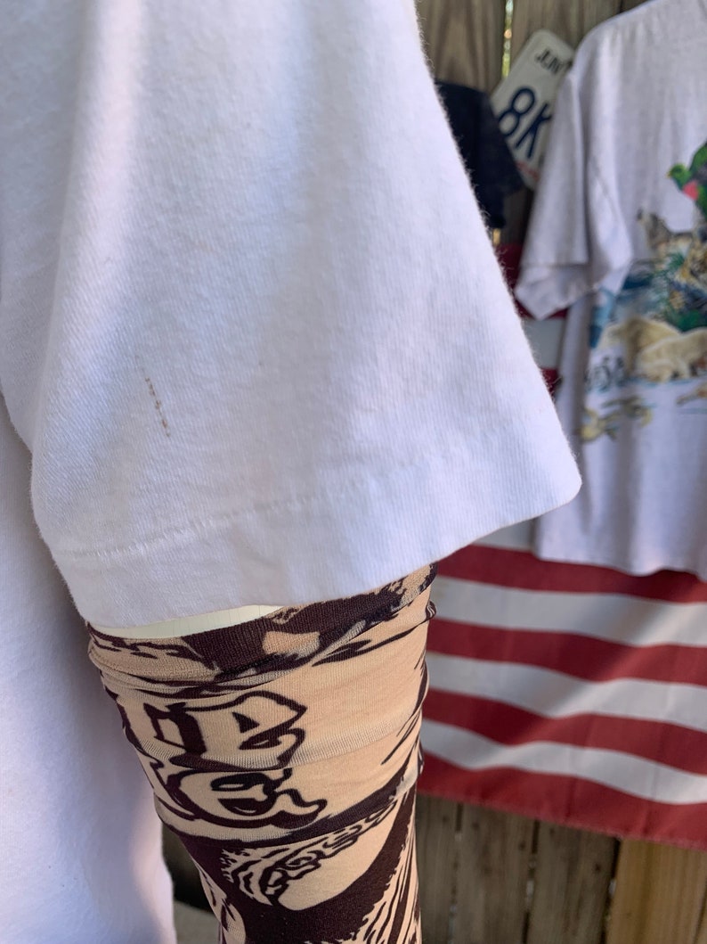 May include: White t-shirt with a brown stain on the sleeve. The sleeve is rolled up and a brown and beige patterned sleeve is visible underneath.
