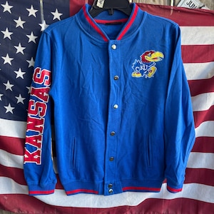 May include: A blue bomber jacket with red and white trim. The left sleeve has the word "KANSAS" in red block letters. The jacket features a patch with a cartoon bird and the letters "KU".