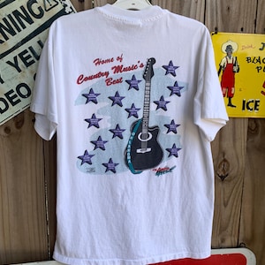 May include: White t-shirt with a graphic of a guitar and stars. The text "Home of Country Music's Best" is printed on the shirt. The graphic also includes the text "Oklahoma".