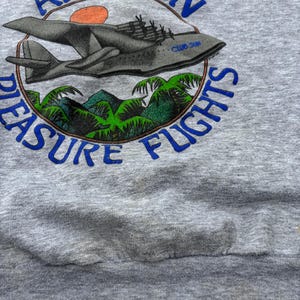 Puede incluir: Sudadera gris con un gr&aacute;fico que presenta un avi&oacute;n vintage, un sol poniente y follaje tropical. El texto "AMAZON PLEASURE FLIGHTS" est&aacute; impreso en azul alrededor del dise&ntilde;o. La sudadera tiene un aspecto desgastado.
