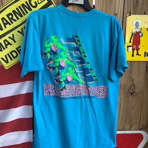 Pode incluir: Uma camiseta azul turquesa com um gráfico de sapos verdes patinando. O texto "Team Bullfrog Amphibious Formula Sunblock" está impresso na camiseta.