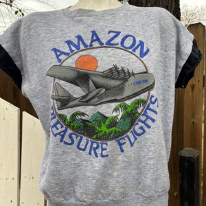 Puede incluir: Sudadera gris de manga corta con un dise&ntilde;o gr&aacute;fico. El dise&ntilde;o presenta un avi&oacute;n gris, un sol naranja y las palabras "AMAZON PLEASURE FLIGHTS" en azul. Las mangas son negras.