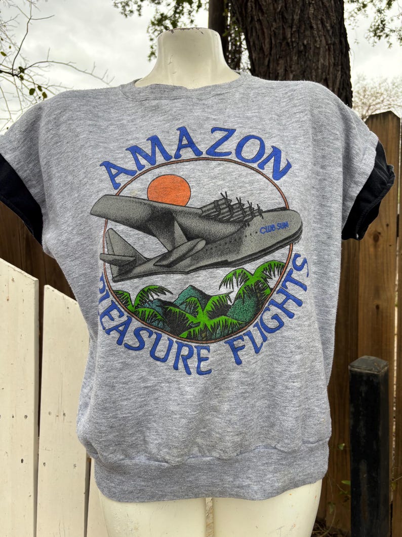 Puede incluir: Sudadera gris de manga corta con un dise&ntilde;o gr&aacute;fico. El dise&ntilde;o presenta un avi&oacute;n gris, un sol naranja y las palabras "AMAZON PLEASURE FLIGHTS" en azul. Las mangas son negras.