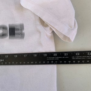 May include: White t-shirt with a black and gray graphic print. The graphic is a stylized image of a rectangle with a smaller rectangle inside. The shirt is measured with a ruler, showing the length is 50 cm.