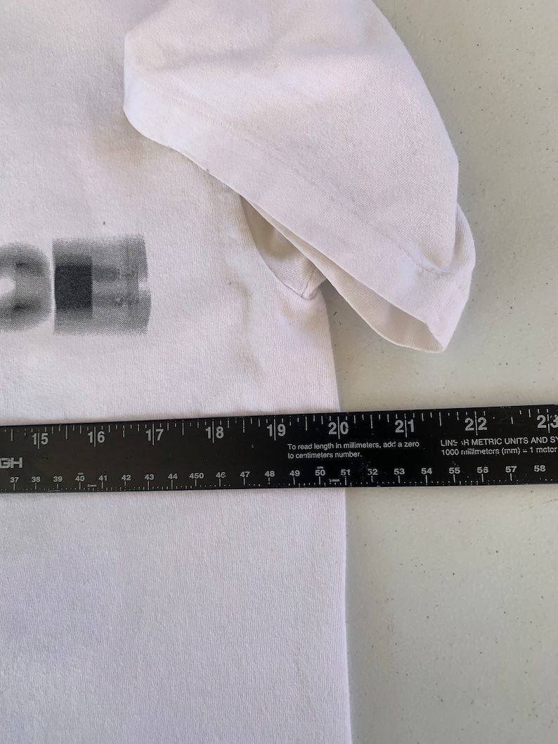 May include: White t-shirt with a black and gray graphic print. The graphic is a stylized image of a rectangle with a smaller rectangle inside. The shirt is measured with a ruler, showing the length is 50 cm.