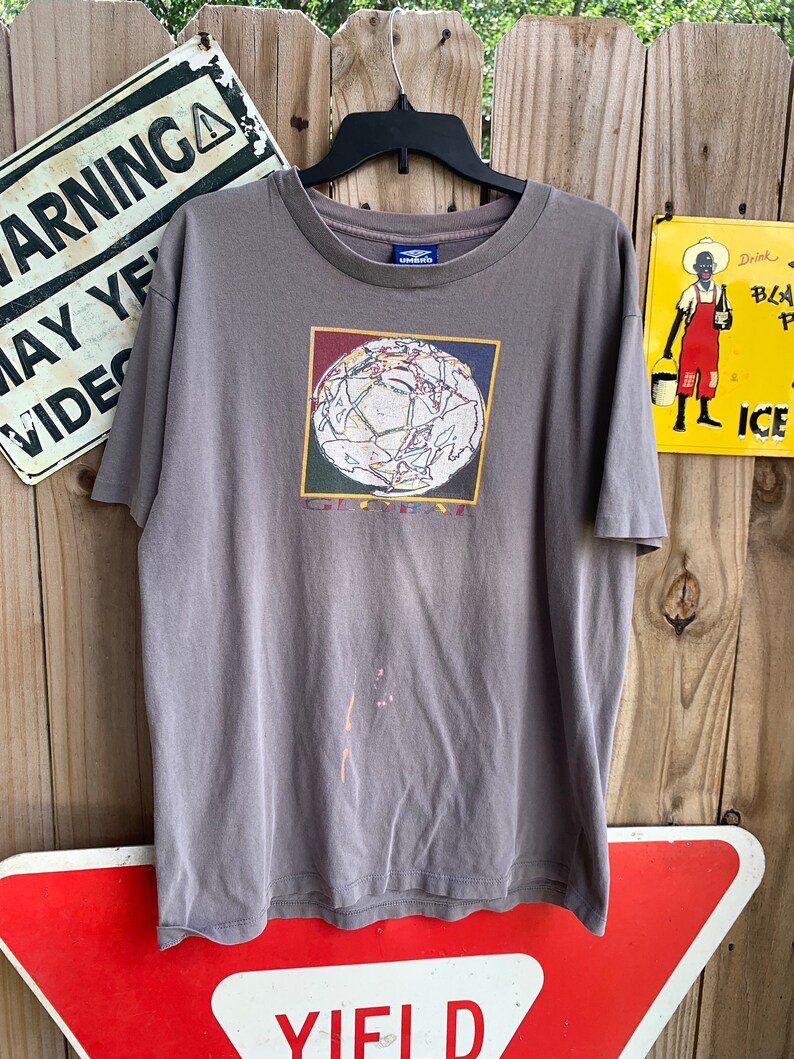 May include: A grey t-shirt featuring a graphic of a colourful globe with the word "Global" printed beneath it. The shirt has a crew neck and short sleeves. The brand name "Umbro" is printed on the tag.