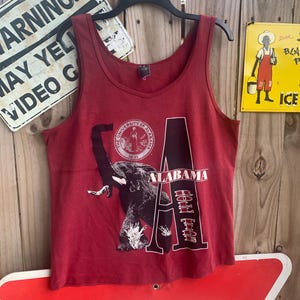 May include: A maroon tank top with a large white letter "A" printed on the front. The letter "A" has the words "Alabama" and "Roll Tide" printed inside it. An elephant is printed behind the letter "A".