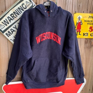 May include: A navy blue hooded sweatshirt with a red "WISCONSIN" logo on the front. The sweatshirt is hanging on a hanger in front of a wooden fence.