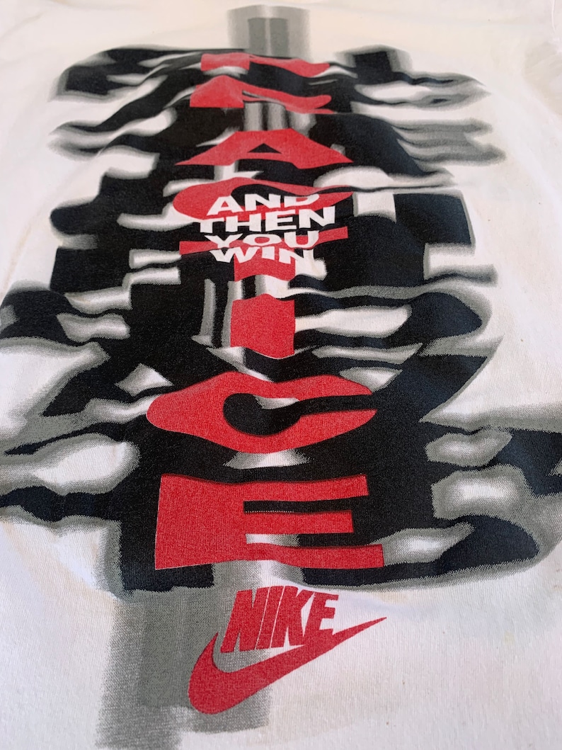 May include: White t-shirt with a black and white graphic design of the word "JUST DO IT" with the words "AND THEN YOU WIN" in red. The Nike swoosh logo is in red at the bottom.