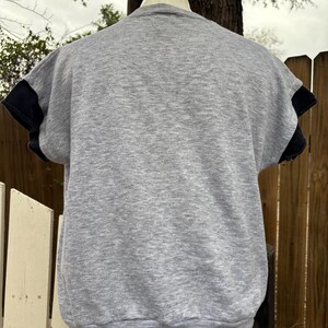Puede incluir: Una sudadera gris de manga corta con mangas negras. La parte trasera de la camisa es visible, mostrando el cuello redondo y las mangas cortas. La camisa se muestra en un maniqu&iacute;.