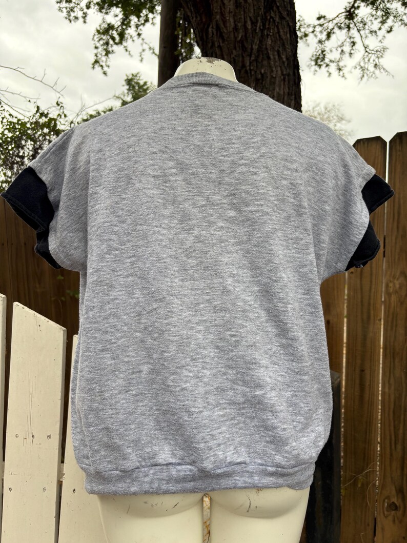 Puede incluir: Una sudadera gris de manga corta con mangas negras. La parte trasera de la camisa es visible, mostrando el cuello redondo y las mangas cortas. La camisa se muestra en un maniqu&iacute;.